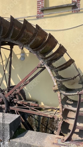 Antique Water Wheel Mechanism: A close-up view of an old, rusty water wheel, its wooden paddles worn by time and use, set against a textured wall. Robbiano, Italy