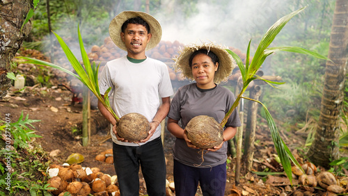 Two young asian farmers holding sprouted coconut seedlings, copra smoke drying on bamboo racks in background, smiling portrait, indonesian people, flores island, indonesia, southeast asia