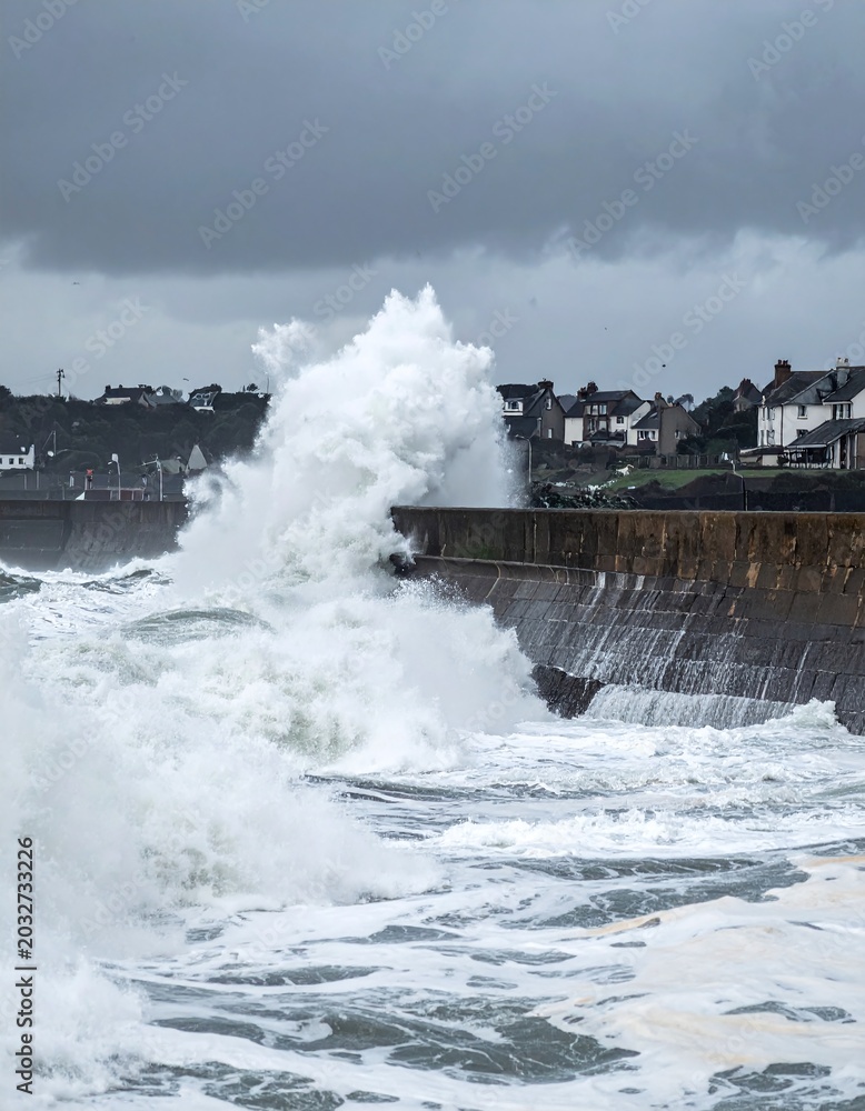 Obraz premium Powerful ocean waves crash against a stone seawall under a stormy sky, highlighting natures strength and coastal vulnerability. Ideal for environmental, climate, and travel safety content.