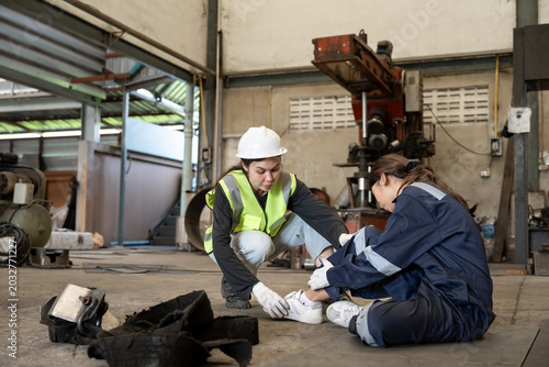 Young Asia female engineer with hurting knee sitting on the floor and female worker in safety helmet helping and calling ambulance