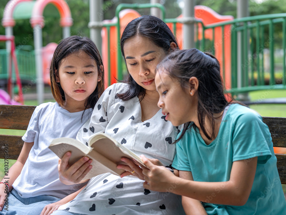 Obraz premium Asian mother reading a book to her two young daughters while sitting on a bench in the park.