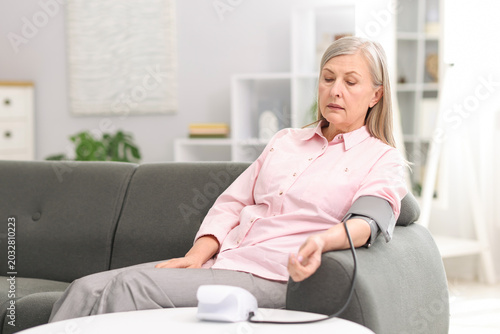 Senior woman measuring blood pressure on sofa at white table indoors