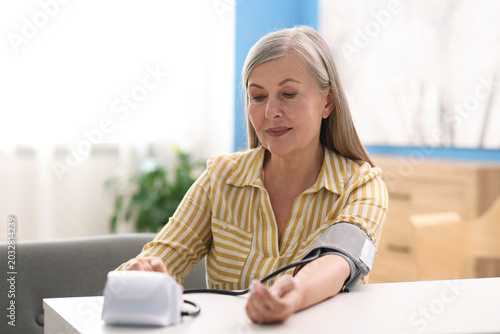 Senior woman measuring blood pressure at white table indoors