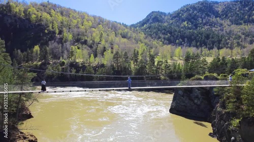 Suspension Bridge Over Muddy River Two Hikers Traverse Narrow Walkway Across Fastflowing Brown River Framed By Forested Foothills And Sunlit Mountains Drone View Captures Cables, Rocky Cliffs, Spring