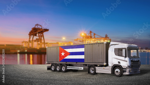 Photography Cuban Cargo Truck at Port: A modern truck carrying a shipping container adorned with the Cuban flag drives along a port at sunset, with cranes and illuminated harbor in the background