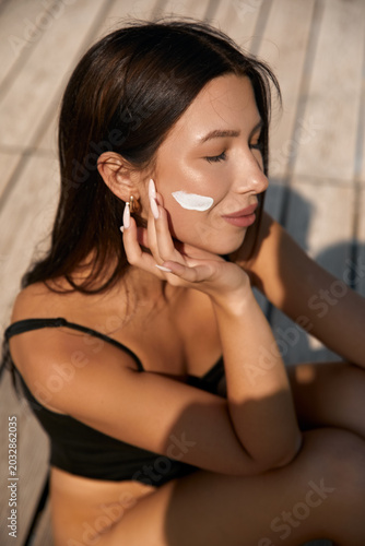Quadro su tela Young woman applies sun protection cream to her cheek while relaxing outdoors on