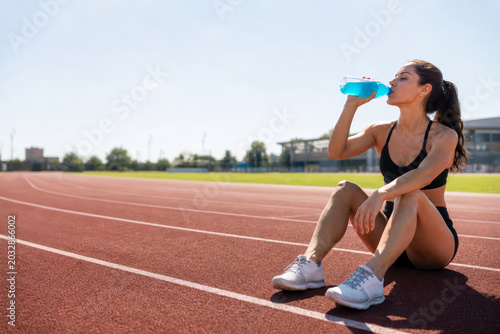 Fit woman in athletic wear drinks from a blue sports drink bottle while sitting on a running track.
