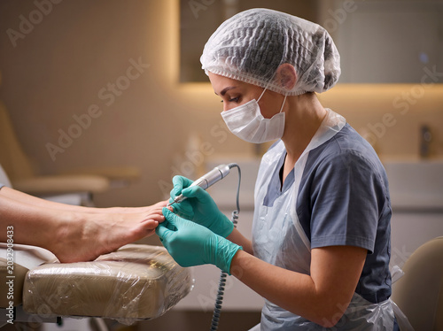 Female podiatrist in face mask performing hardware pedicure in clinic