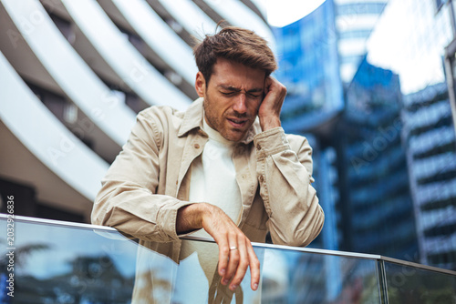 Young Man Holding Head in Pain Leaning on Glass Railing Outside Modern Office Buildings