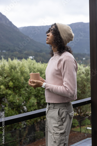 Non-binary adult standing on wooden balcony holding ceramic mug and gazing toward distant mountains