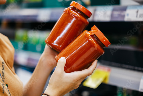 Person holding two jars of tomato sauce with orange lids while shopping in a grocery store aisle, shelves filled with various products in the background