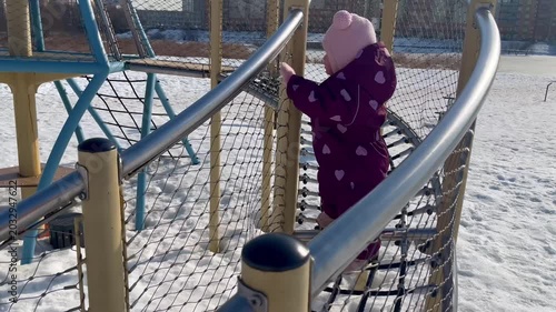 Young girl in purple snowsuit climbs on playground netting structure, navigating the ropes with determination while surrounded by snow-covered ground and playground equipment in a winter setting