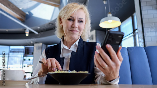 Caucasian Elderly Businesswoman Checking Calendar On Smartphone During Lunch At Modern Cafe, Concentrated Expression, Fork At Plate, Digital Multitasking Between Messages And Meal, Efficient Midday