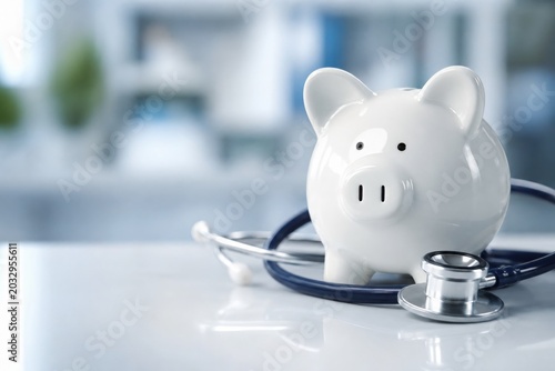 A piggy bank with a stethoscope on a desk in a clinical setting, symbolizing the cost of healthcare and medical savings.