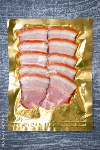 Vacuum sealed sliced smoked bacon in gold foil packaging on rustic wooden grey table surface.