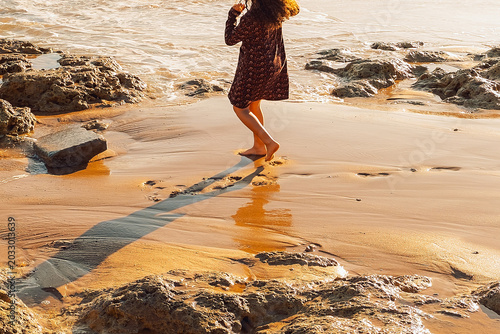 young Child plays by ocean at sunset, exploring rocks  along shore while waves crash nearby. girl walks along beach, enjoying feeling of sand and water.  sun sets in background,  warm atmosphere.