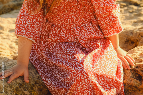 Girl in  red dress stands on beach by ocean during sunset . child stands on large rocks by sea, crossing her arms. sun sets in background, warm glow over scene. waves crash gently against the shore.