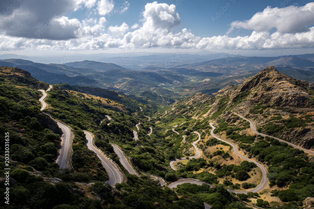 Obraz premium Aerial view of a winding mountain road through a lush green forest with countless switchbacks, descending towards a misty coastal landscape.