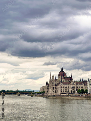 View of the Hungarian Parliament Building on the banks of the Danube River under dramatic cloudy skies. Iconic European landmark and travel destination in Budapest, Hungary.