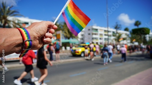 Photos Person's hand displaying a colorful rainbow wristband and waving a small rainbow