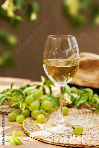 White wine glass on wicker placemat with green grapes, vine leaves and straw hat on wooden table