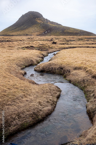 vertical fotography of river flows through a dry grassland with a mountain in the background on the Reykjanes Peninsula in Iceland during a cold March day.