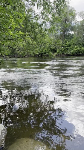 Tranquil river water surface with a beautiful sky and green tree reflection. Picturesque view of calm river flow on a cloudy spring day. Peaceful nature and water backdrop