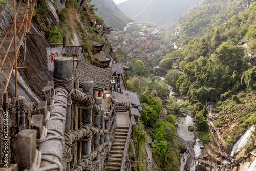 Tableau sur toile Wangxian Valley, china - Ancient cliff-side village with wooden railings, stone steps, traditional rooftops, and a river gorge cutting through lush green mountains