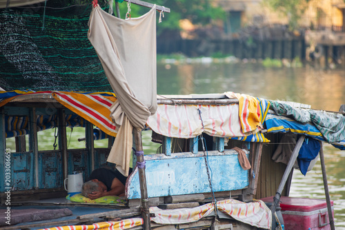Konstfotografi A senior man resting and sleeping inside a weathered, traditional wooden boat docked along a river canal in Vietnam