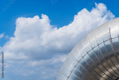 Принти на полотні Close-up of a large silver metallic insulation pipe elbow at a petrochemical plant against a bright blue sky with white clouds