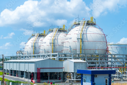 A row of large industrial spherical storage tanks at a petrochemical plant under a bright blue sky with clouds.