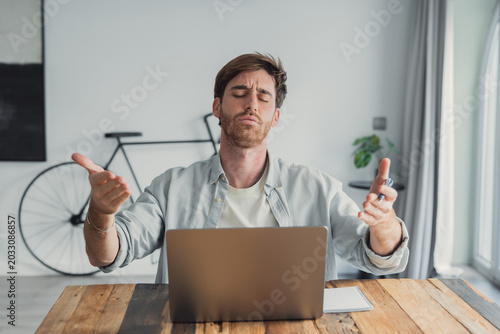 Fotografi Frustrated young adult man sitting at a wooden table with a laptop, expressing s