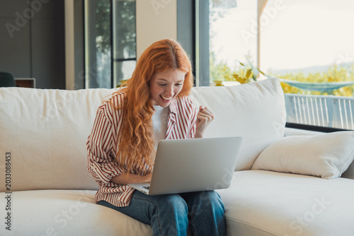 Foto Young redheaded woman on a white couch celebrates good news while working on her