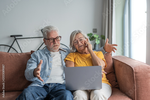 Fotografija Senior couple sitting on a sofa, looking frustrated and confused while trying to
