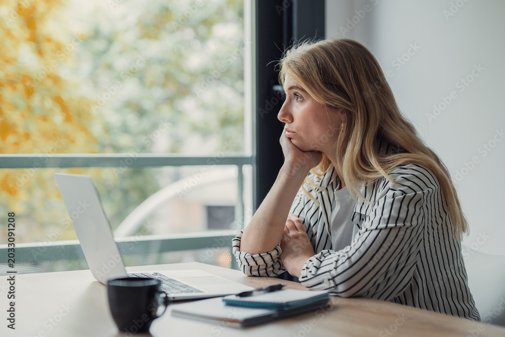 Fototapeta premium Young woman in a striped shirt sits at a desk with laptop and coffee, daydreaming and staring out a large window during a distracted, unmotivated work-from-home moment