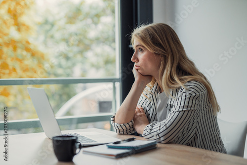 Schilderij op canvas Young woman sitting at a desk with a laptop and coffee, looking out the window w