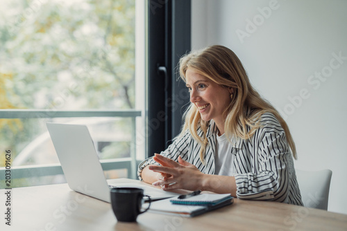 Young adult woman smiling and gesturing during a video call on her laptop, working remotely from her home office with a coffee cup and notebook on the desk
