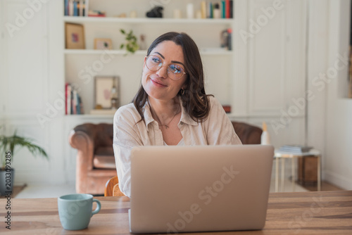 Fotografie Young professional woman in glasses sitting at a wooden table with laptop and co