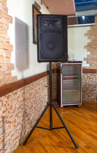 stone wall background, perfect for events, concerts, parties and public speaking setups. Professional large black audio speaker mounted on a sturdy metal tripod stand indoors with a wooden floor .
