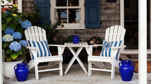White adirondack chairs and table on wooden porch with blue hydrangeas and pots. Coastal cottage outdoor seating area with blue striped cushions and bright blue vases.