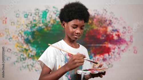 African boy standing at colorful stained wall while holding brush and point at camera. Happy high school student wearing white shirt with stain color and look at camera with confident. Edification