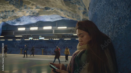 A contemporary underground station setting featuring travelers engaged with their mobile devices. The space is characterized by unique architectural elements and vibrant lighting that enhances the com