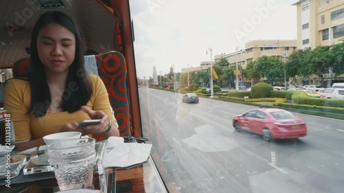 A woman sits on a bus, savoring a refreshing drink while enjoying a scenic view of the city. The urban landscape features green trees and bustling streets, creating a vibrant atmosphere.