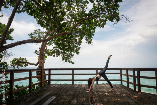 Photography Peaceful tropical wellness scene featuring a woman practicing yoga on a wooden oceanfront deck surrounded by lush green trees and overlooking the sea