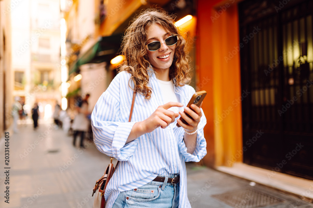 Fototapeta premium A young woman wearing sunglasses and holding a phone strolls along the sunny streets of the city. A smiling woman poses with a phone outdoors. Concept of style, walking, and technology.