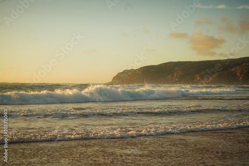Dramatic sunset over the Atlantic Ocean in Portugal, with powerful waves crashing onto the shore and rolling coastal hills fading into the distance under a glowing evening sky.