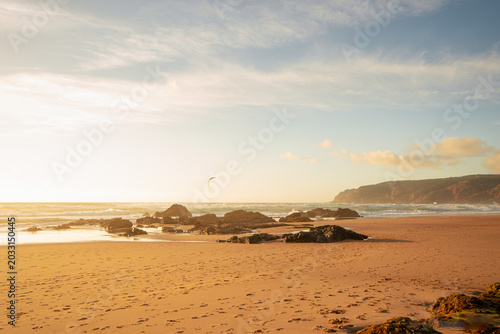 Golden sunset over the Atlantic Ocean with black volcanic stones scattered along the beach, while a kiteboarder rides in the distant horizon under a glowing sky, capturing the energy of wind and sea.