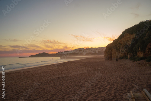 Golden sunset over a misty sandy beach in Portugal’s Algarve region, with soft pink clouds in the sky, a hazy ocean horizon, and distant coastal hills dotted with houses glowing in warm evening light.