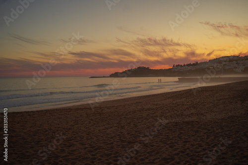 Golden sunset over a misty sandy beach in Portugal’s Algarve region, with soft pink clouds in the sky, a hazy ocean horizon, and distant coastal hills dotted with houses glowing in warm evening light.