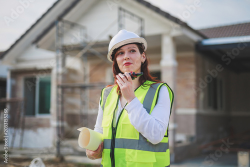 Fotografi Female engineer wearing safety helmet and vest, holding blueprint, communicating
